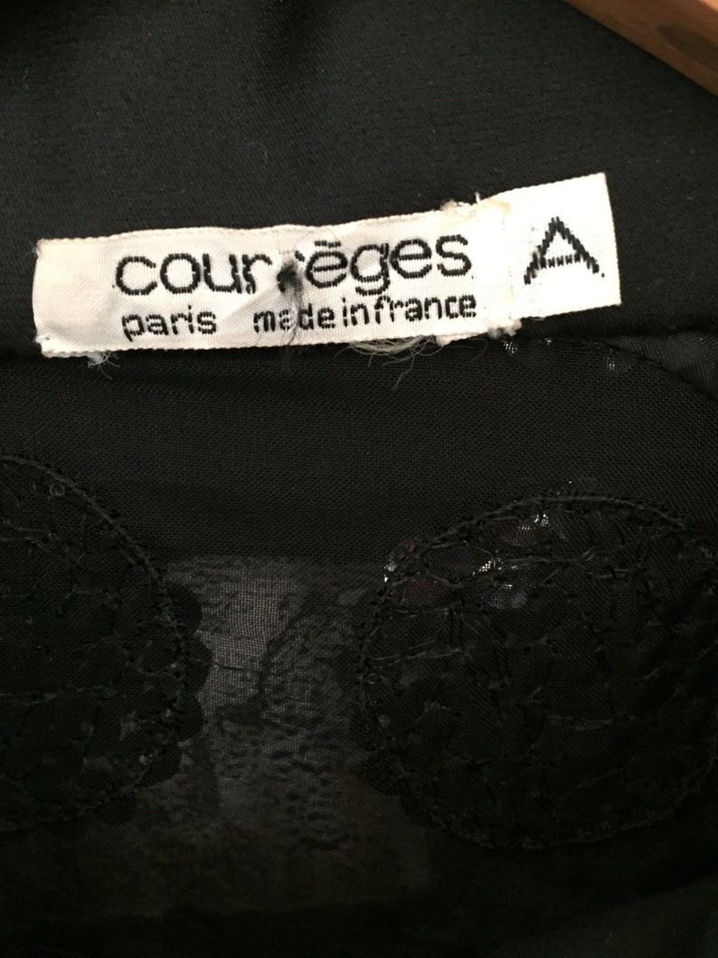 Op de afbeelding: Zwart doorschijnend topje met een zwarte kanten overlay en een wit label met de tekst "Courr&egrave;ges Paris made in France" met een hoofdletter "A" gedrukt aan de rechterkant van het label.