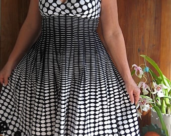 May include: A woman wearing a black and white polka dot dress with a skull print hem. She is wearing sunglasses and a beaded necklace.