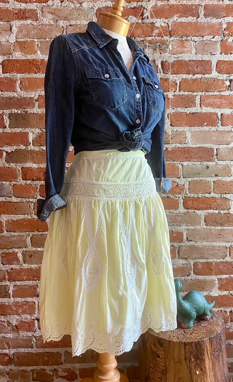 May include: A yellow cotton skirt with white embroidery and lace trim. The skirt is gathered at the waist and has a full, flowing silhouette.