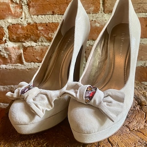 Puede incluir: Un par de tacones de plataforma de color gris claro con un detalle de lazo y adornos de piedras preciosas. Los zapatos son de la marca "BCBGMAXAZRIA".