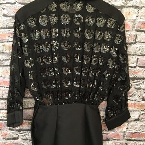 Op de afbeelding: Zwart shirt met pailletten, een kraag en lange mouwen. Het shirt heeft een getailleerd bovenlichaam en een losse, vloeiende rok.