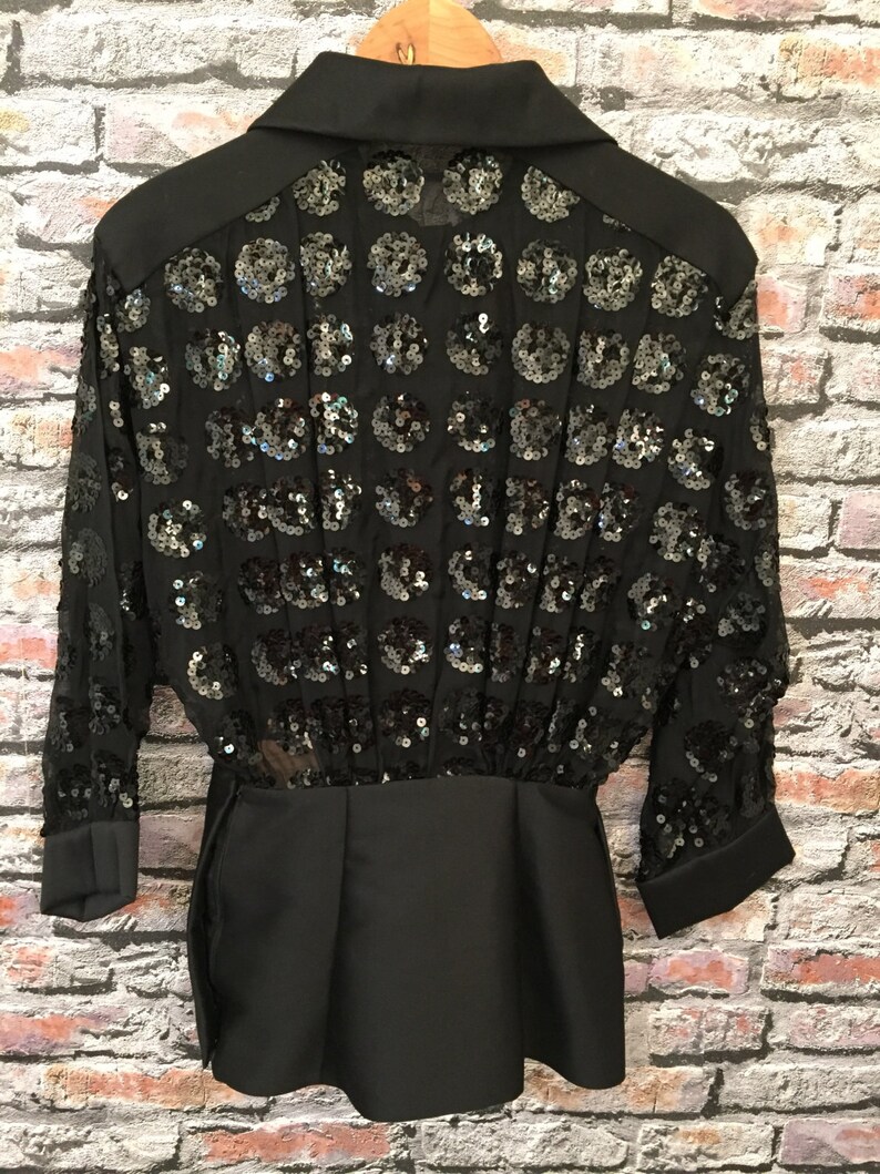 Op de afbeelding: Zwart shirt met pailletten, een kraag en lange mouwen. Het shirt heeft een getailleerd bovenlichaam en een losse, vloeiende rok.