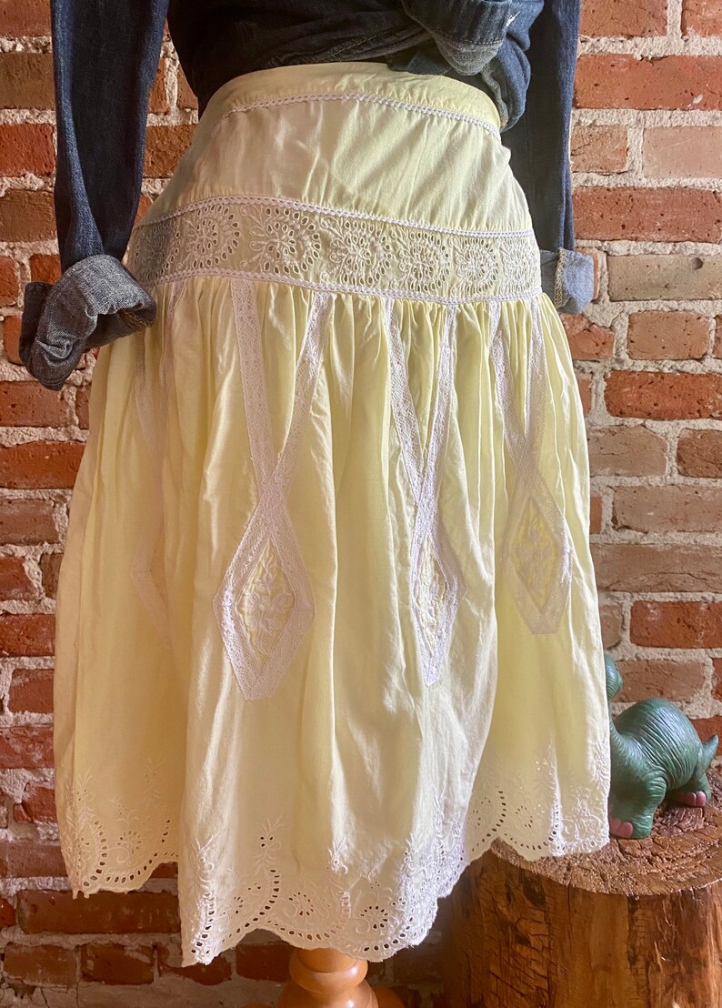 May include: A pale yellow cotton skirt with white eyelet lace trim. The skirt has a gathered waist and a full, flowing silhouette.