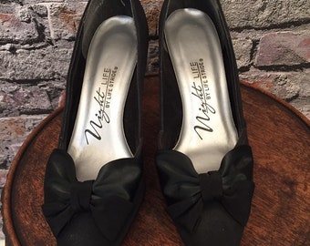 May include: A pair of black satin pumps with a bow on the toe. The shoes are labeled "Night Life by Life Stride".