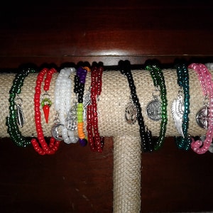 May include: A collection of beaded bracelets in various colors, including green, red, white, and pink, displayed on a tan fabric-covered stand. Each bracelet features unique charms, such as feathers, chili peppers, and compasses. The bracelets are made with small beads.