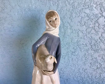May include: A porcelain figurine of a girl in a blue dress and white head scarf holding a white lamb.