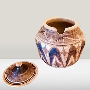 May include: A brown and white ceramic jar with a lid. The jar has a geometric pattern with brown and blue accents. The lid has a brown and white striped pattern.