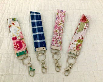 May include: Four fabric key fobs with metal hardware. The patterns include floral, plaid, and patchwork designs in pink, blue, and green. Each fob has a metal ring and clasp for attaching keys.