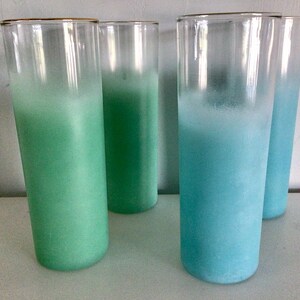 May include: Four tall, clear drinking glasses with gold rims. The glasses are filled with a gradient of color, transitioning from green to blue. The glasses are cylindrical and appear to be made of glass.