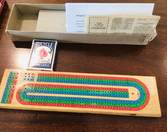 May include: A wooden cribbage board with red, green, and blue tracks, a deck of Bicycle playing cards, and a box containing instructions. The board has a curved design and is ready for a game of cribbage.