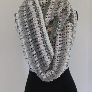 Puede incluir: Una bufanda infinita de crochet gris y blanco con un patrón texturizado. La bufanda se muestra en un maniquí negro.