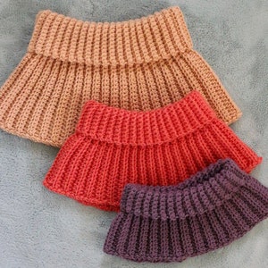 Pode incluir: Três cachecóis de crochê em tamanhos e cores diferentes. O maior é castanho claro, o do meio é cor de coral brilhante e o menor é roxo escuro.