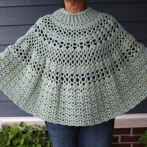 Puede incluir: Un poncho de ganchillo en un esquema de color azul claro y verde. El poncho tiene un escote amplio y abierto y un ajuste holgado y fluido.