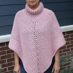 May include: A pink crocheted poncho with a high neck collar. The poncho is worn by a person with blue jeans.