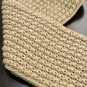 Può includere: Un primo piano di una sciarpa all'uncinetto di colore marrone chiaro. La sciarpa è realizzata con un semplice motivo a punto e ha un aspetto testurizzato.