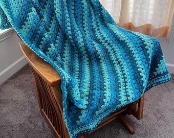 Peut inclure: Une couverture en crochet bleu et vert drapé sur un fauteuil à bascule en bois. La couverture a un motif rayé et est faite avec un fil doux.