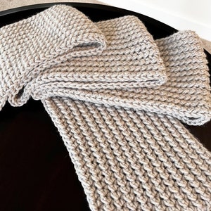 May include: A light gray knitted scarf and matching square items, possibly washcloths or coasters, are arranged on a dark surface. The items have a textured, woven appearance, suggesting a handmade or artisanal quality. The neutral color palette makes them versatile.