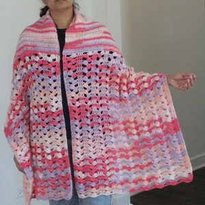 May include: A crocheted shawl in shades of pink, purple, and white. The shawl has a lacy pattern and is draped over a person's shoulders.