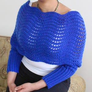 Może przedstawiać: Sweter w kolorze królewskiego błękitu wykonany na szydełku z szerokim, otwartym dekoltem i długimi rękawami. Sweter ma delikatny, ażurowy wzór.