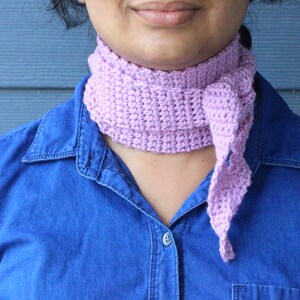 May include: A lilac crochet scarf is worn by a person in a blue denim shirt. The scarf, made from soft yarn, has a textured appearance and a twisted, knotted design. The shirt has a buttoned collar.