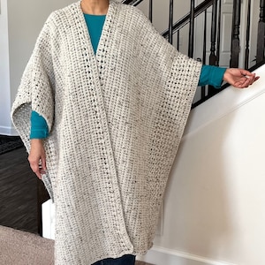 May include: A long, off-white crocheted shawl with a textured pattern. The shawl is open in the front and has a wide, flowing shape.