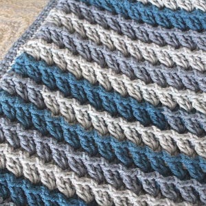 May include: A close-up of a crocheted blanket with a striped pattern in shades of blue and grey.