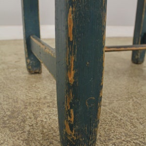Op de afbeelding: Close-up van een houten kruk met een donkerblauwe teal verf afwerking. De verf is gechipt en versleten, waardoor het natuurlijke hout eronder zichtbaar wordt. De kruk heeft een eenvoudig, rustiek ontwerp.