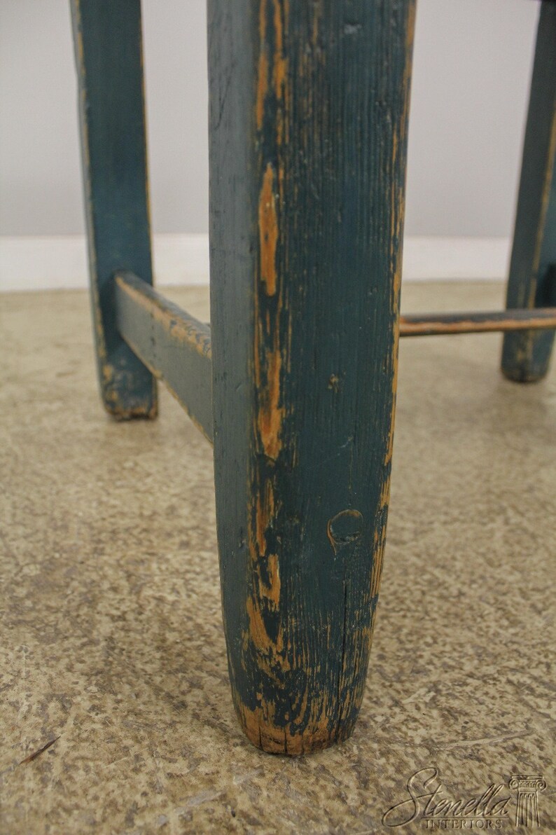 Op de afbeelding: Close-up van een houten kruk met een donkerblauwe teal verf afwerking. De verf is gechipt en versleten, waardoor het natuurlijke hout eronder zichtbaar wordt. De kruk heeft een eenvoudig, rustiek ontwerp.
