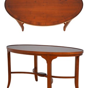 May include: An oval wooden table with a smooth, polished brown surface featuring a dark border inlay. The table is supported by four curved legs connected by a central cross-stretcher base with a small, rounded wooden accent piece.