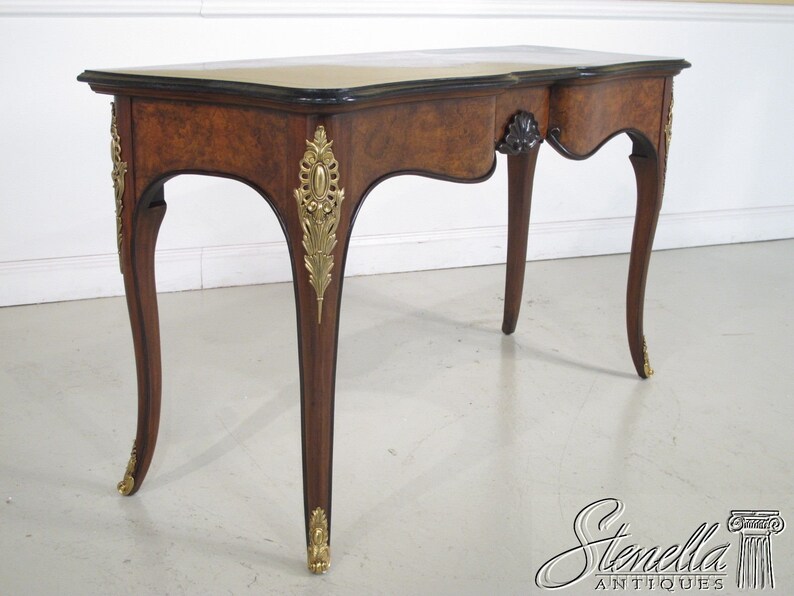 May include: A brown wooden writing desk with a black top and gold accents. The desk has a single drawer and curved legs.