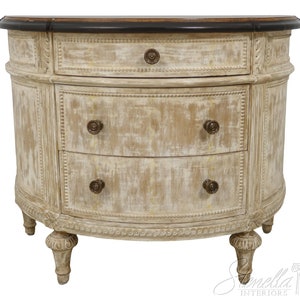 May include: A white, distressed, oval-shaped chest of drawers with three drawers and a dark wood top. The chest has ornate carvings and brass knobs.