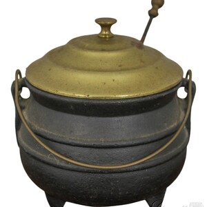 May include: A black cast iron pot with a brass lid and a wooden handle. The pot has three legs and a single handle.