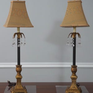 May include: Two matching table lamps with gold-colored bases and black stems. The lampshades are a light tan color with a patterned design. Each lamp has crystal accents hanging from the top. The lamps are placed on a wooden surface.