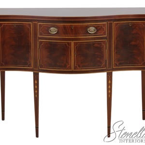 May include: A mahogany-colored sideboard with a curved front, featuring two doors and a central drawer with brass hardware. The piece has slender, tapered legs with decorative inlays, and is suitable for a dining room or living room.