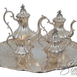 May include: A silver tea set on a silver tray. The set includes a teapot, creamer, sugar bowl, and a serving tray with handles. The pieces have ornate details and are reflective. The set is likely vintage or antique.
