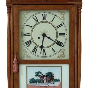 May include: A wooden wall clock with a white face and black Roman numeral hour markers. The clock has a small picture of a house below the clock face. The clock has a decorative top with three gold finials.