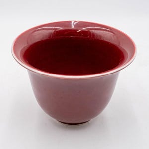 May include: A deep red ceramic bowl with a smooth, glossy finish and a wide, flared rim. The interior and exterior feature a consistent dark crimson glaze, while the thin edge of the rim shows a subtle white line, highlighting the bowl's shape.