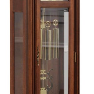 May include: A tall wooden grandfather clock with a rich brown finish, featuring an arched top, a glass-fronted door, and a gold-toned clock face with Roman numerals. Inside, three brass weights and a large circular brass pendulum are visible.