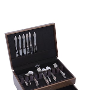May include: A silver flatware set with ornate floral detailing, displayed in a wooden case with a black velvet lining. The set includes knives, forks, and spoons. A small pouch is also included. The case is open, revealing the arrangement of the silverware.