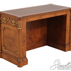 May include: A rectangular wooden desk featuring a rich burl wood grain finish, accented with ornate gold-toned metal floral carvings and fluted column details. The desk surface is framed by a decorative border, and the base includes a logo reading Stenella Interiors.