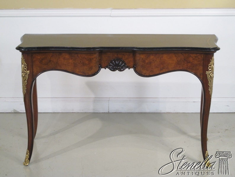 May include: A brown wooden console table with a black marble top and gold accents. The table has a curved front and legs, and features a decorative carved design on the front.