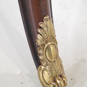 May include: Close-up of a wooden table leg with a gold metal decorative foot. The foot features a stylized floral design.
