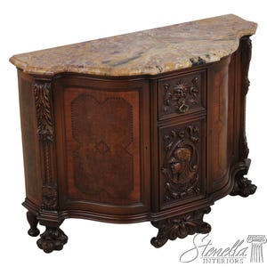 May include: A dark wood sideboard featuring a curved front, a mottled beige and brown marble top, and intricate carvings. The cabinet includes a central drawer with a metal pull, a door panel with a relief portrait, and four carved claw feet.
