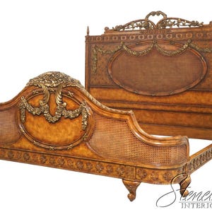 May include: A wooden bed frame featuring a tall headboard and a lower footboard, both with oval cane panels, intricate gold-toned floral carvings, and swags. The frame has a warm brown finish, carved detailing along the base, and short feet.