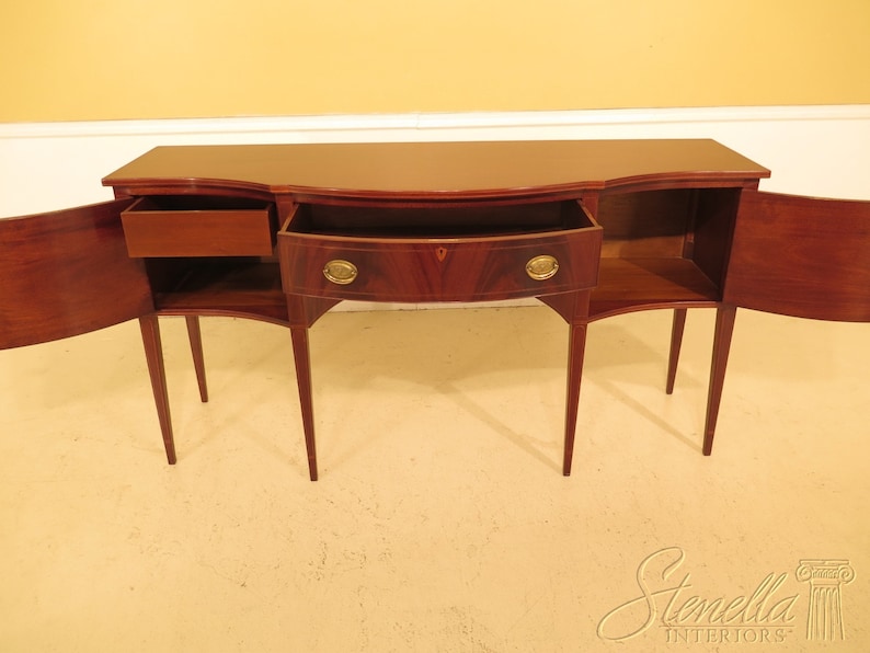 Pu&ograve; includere: Un credenza in legno di mogano con tre cassetti e due ante. Le ante sono aperte, rivelando gli scaffali interni. La credenza ha un fronte curvo e quattro gambe.