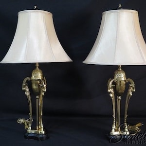 May include: A pair of gold-toned table lamps with white fabric lampshades. The lamps feature a decorative base with a stylized design.