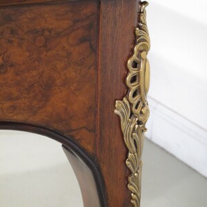 May include: A close-up of a wooden table leg with a decorative brass accent. The table leg is curved and has a dark brown finish. The brass accent is ornate and features a floral design.
