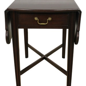 May include: A dark brown wooden drop-leaf table with a single drawer and brass hardware. The table has a cross-shaped base and is supported by four legs.