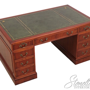 May include: A large, wooden desk with a green leather top and multiple drawers. The desk is a rich, reddish-brown color with brass hardware. The desk measures approximately 60 inches wide and 30 inches deep.