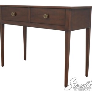 May include: A dark brown wooden console table featuring two rectangular drawers with round brass-toned knobs. The table stands on four tapered legs and has a flat rectangular top surface. The Stenella Interiors logo is visible in the corner.